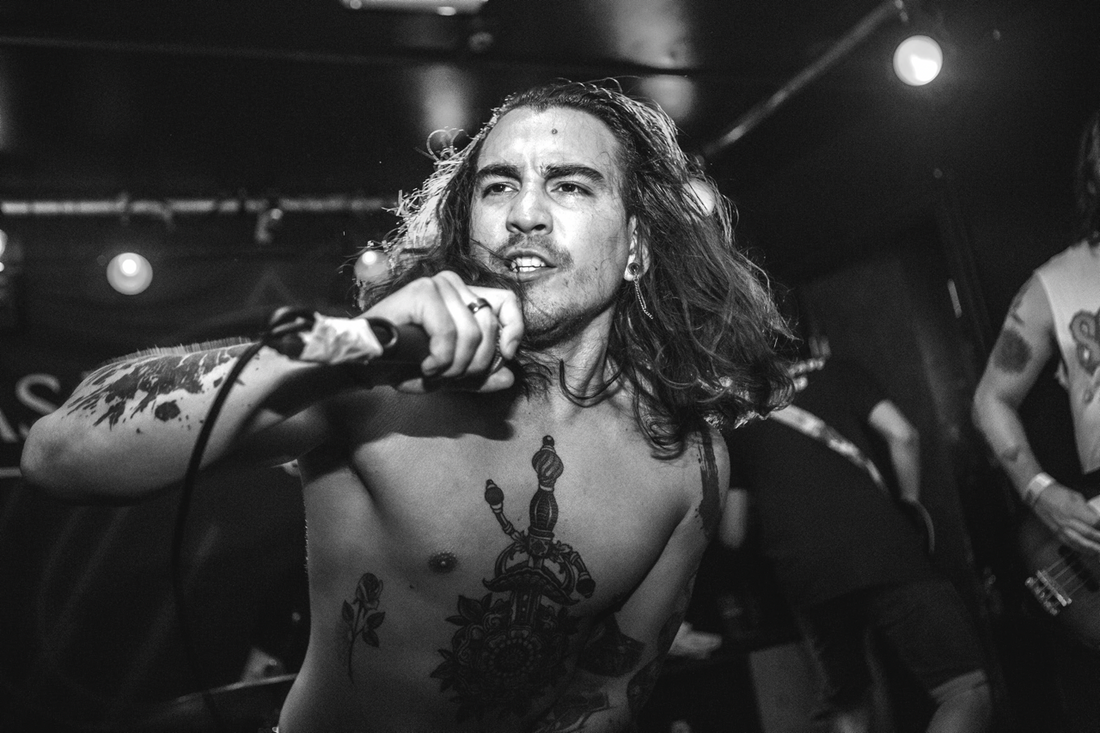 Shirtless rock singer with long wavy black hair and tattoos performing on stage with microphone in hand.
